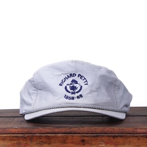 May include: A light gray baseball cap with a curved brim. The cap features the text "RICHARD PETTY" in a circular design with the years "1958-88" below. The cap is sitting on a wooden surface.