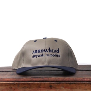 Könnte beinhalten: Beige Baseballkappe mit marineblauem Schirm und dem gestickten Text "ARROWHEAD drywall supplies" in Marineblau. Die Kappe liegt auf einer Holzoberfläche.