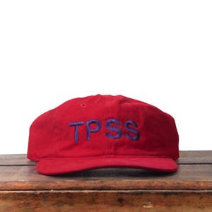 Puede incluir: Gorra de béisbol roja con las letras bordadas "TPSS" en azul. La gorra está hecha de un material suave y descansa sobre una superficie de madera. El sombrero tiene una visera curva y un estilo clásico e informal.