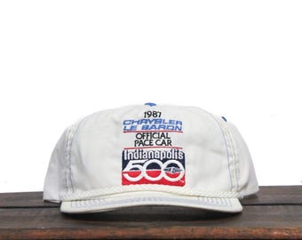 Könnte beinhalten: Weiße Baseballkappe mit blauem Besatz. Die Vorderseite der Kappe zeigt ein gesticktes Emblem mit dem Text "1987 Chrysler Le Baron Official Pace Car Indianapolis 500".
