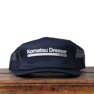 May include: Navy blue trucker hat with a mesh back and a curved brim. The front of the hat features the white text "Komatsu Dresser Company" in a bold, sans-serif font.