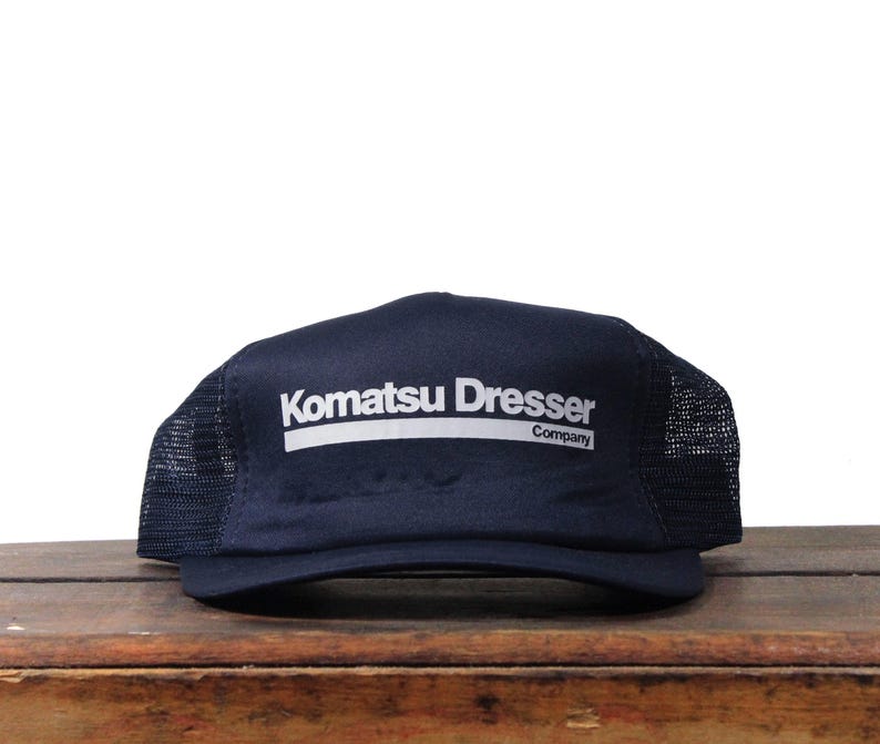 May include: Navy blue trucker hat with a mesh back and a curved brim. The front of the hat features the white text "Komatsu Dresser Company" in a bold, sans-serif font.
