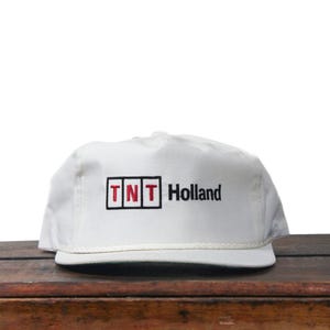 May include: White trucker hat with the text "TNT Holland" in black and red. The hat has a rope detail along the brim and a white mesh back. The hat is sitting on a wooden surface.