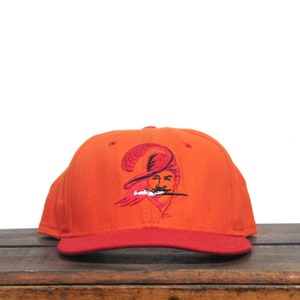 May include: Orange and red baseball cap with an embroidered logo of a pirate wearing a feathered hat and holding a sword. The logo is in red and black.