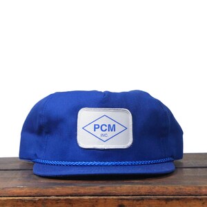 May include: A royal blue baseball cap with a white patch featuring the text "PCM INC." in a diamond shape. The cap has a blue rope detail along the brim and is set against a wooden surface.