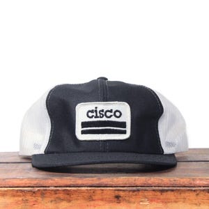 May include: Black and white trucker hat with the word "cisco" on a rectangular patch. The hat has a black front panel, a white mesh back, and a curved brim. The patch has a white background with black text.