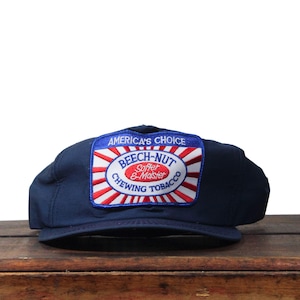 Könnte beinhalten: Marineblaue Baseballkappe mit einem rechteckigen Aufnäher mit der Aufschrift "America's Choice" und "Beech-Nut Softer & Moister Chewing Tobacco". Der Aufnäher hat ein rot-weiß-blaues Design.