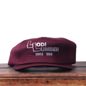 May include: A maroon cap with a flat brim. The cap features the text "LODI LUMBER SINCE 1889" in silver lettering. The cap is sitting on a wooden surface.