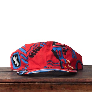 May include: A red baseball cap with a vibrant, abstract pattern in blue, black, and white. The cap is made of fabric and sits on a wooden surface. The brim features a blue and white design.
