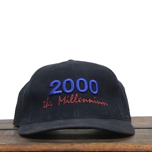 Könnte beinhalten: Marineblaue Baseballkappe mit dem Text "2000 the Millennium" in blauen und roten Fäden gestickt. Die Kappe liegt auf einer Holzoberfläche.