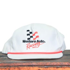 May include: White cap with the text "Western Auto Racing" in black and red. The cap features a checkered flag design and a red rope detail along the brim. The cap is sitting on a wooden surface.