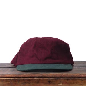 May include: A maroon and green baseball cap. The cap features a maroon crown and a green brim. The cap is made of a soft material and is sitting on a wooden surface.