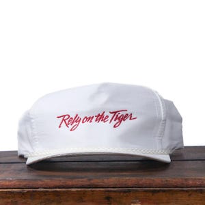 Könnte beinhalten: Weißes Baseball-Cap mit dem rot gestickten Schriftzug "Rely on the Tiger". Die Kappe hat eine Kordel am Rand und liegt auf einer Holzoberfläche.