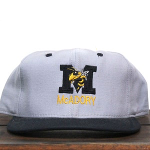May include: A gray and black baseball cap with an embroidered yellow and black bee inside a black letter "M" and the text "MCADORY" below.