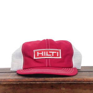 Può includere: Un cappellino trucker rosso e bianco con pannello frontale rosso e la scritta HILTI ricamata in bianco all'interno di un bordo rettangolare. Il cappellino presenta una visiera curva con cuciture bianche e pannelli laterali in rete bianca.