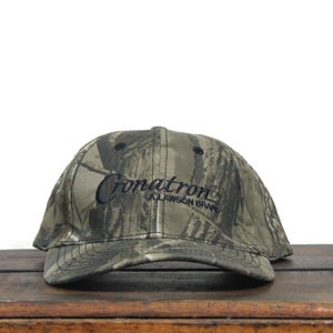 May include: Camouflage baseball cap with the embroidered text "Cronatron A Lawson Brand" in black. The cap features a curved brim and a classic six-panel design. The hat is made of a durable fabric and is suitable for outdoor activities.