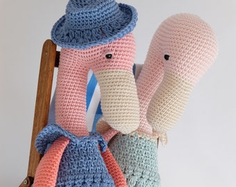 May include: Two crocheted flamingo toys sitting on a wooden chair. One flamingo is wearing a blue hat and a blue dress. The other flamingo is wearing a light blue dress.