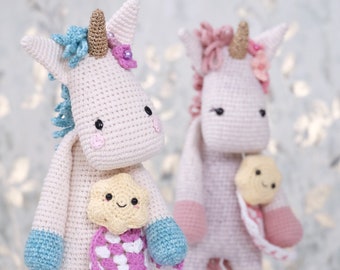 May include: Two handmade crochet unicorns. One is cream-colored with blue and purple accents, a gold horn, and a smiling cloud-shaped pocket. The other is pink with a gold horn and a smiling star-shaped pocket. Both have black eyes.