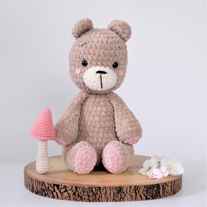 Puede incluir: Un oso de peluche hecho a mano, de color marrón suave, con un hocico blanco y detalles rosas en las patas y mejillas. Una seta rosa y pequeñas flores blancas están junto al oso. El oso está sentado sobre una rodaja de madera.