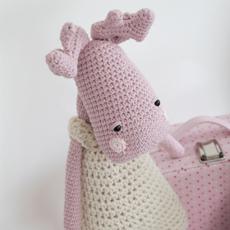 May include: A pink crocheted reindeer toy with a white crocheted dress. The reindeer has black eyes and a pink nose.