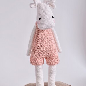 May include: A white crocheted moose wearing pink overalls. The moose has black eyes and pink cheeks. The moose is standing on a wooden log.