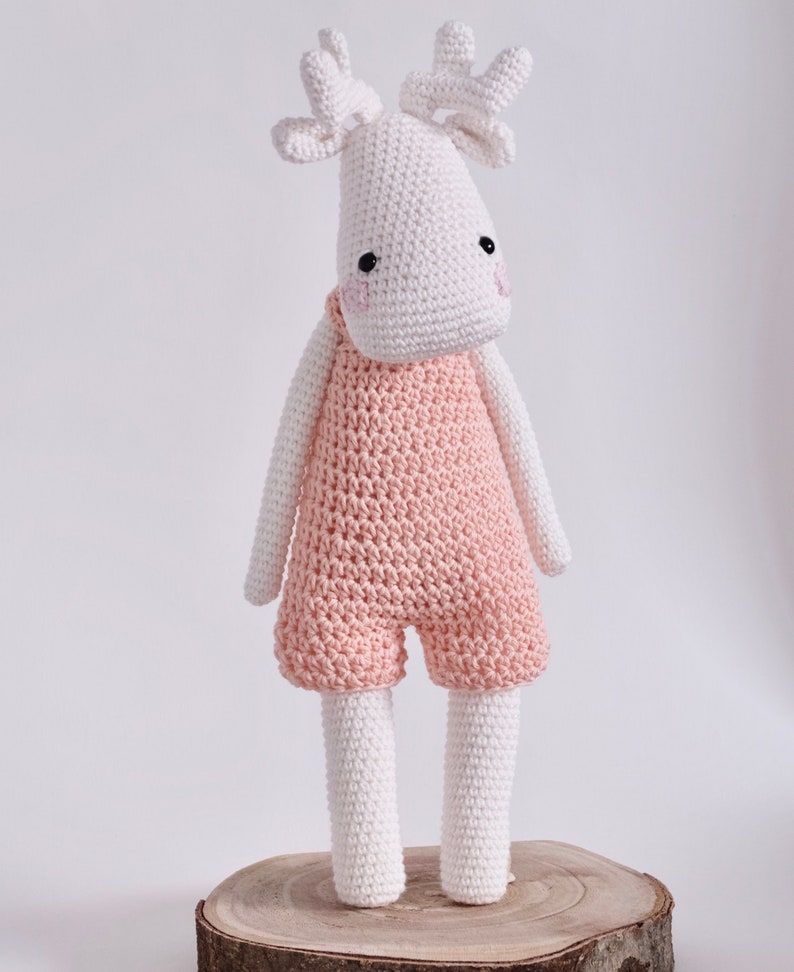May include: A white crocheted moose wearing pink overalls. The moose has black eyes and pink cheeks. The moose is standing on a wooden log.