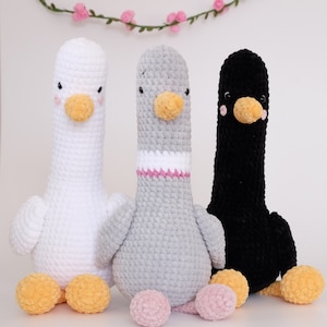 May include: Three crocheted stuffed animals shaped like geese. The geese are white, gray, and black. The white goose has a pink nose and yellow beak. The gray goose has a pink nose and yellow beak. The black goose has a pink nose and yellow beak.