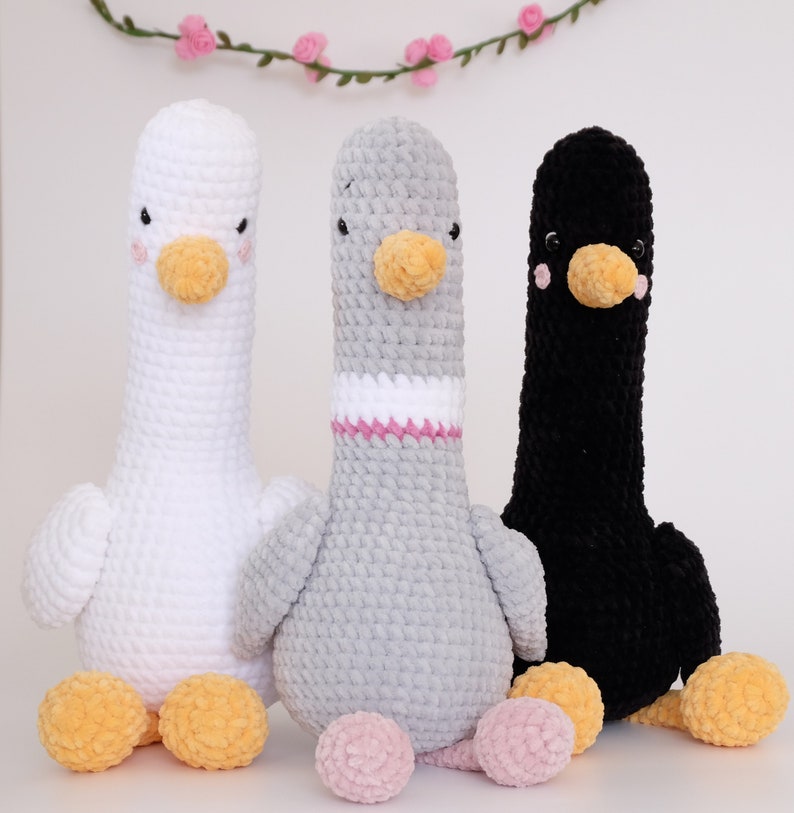 May include: Three crocheted stuffed animals shaped like geese. The geese are white, gray, and black. The white goose has a pink nose and yellow beak. The gray goose has a pink nose and yellow beak. The black goose has a pink nose and yellow beak.