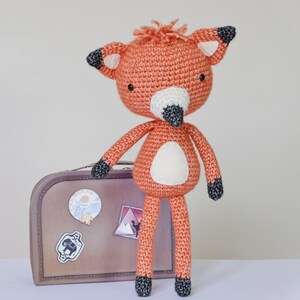 May include: A crocheted orange fox toy with black eyes and a black nose. The fox is standing on a brown suitcase with travel stickers.
