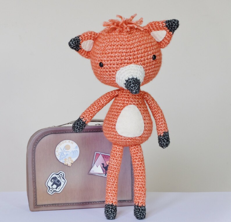 May include: A crocheted orange fox toy with black eyes and a black nose. The fox is standing on a brown suitcase with travel stickers.