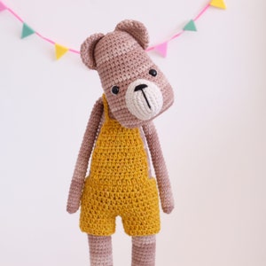 May include: A crocheted stuffed animal bear wearing a yellow crocheted overall. The bear has a brown and pink striped body and a white crocheted nose.