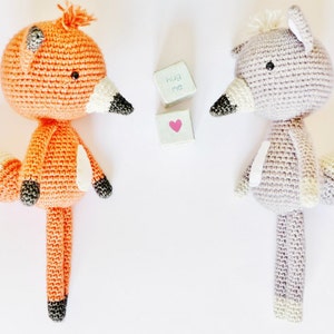 May include: Two crocheted stuffed animals, a fox and a wolf, standing on a white background. The fox is orange with a white tail and black eyes. The wolf is gray with a white tail and black eyes. The text "hug me" is written on a small wooden block.