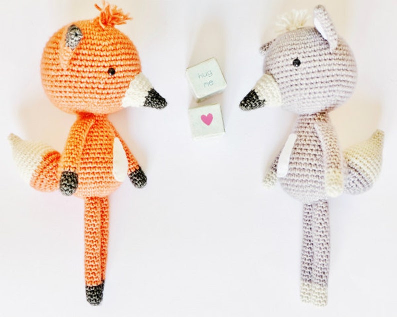 May include: Two crocheted stuffed animals, a fox and a wolf, standing on a white background. The fox is orange with a white tail and black eyes. The wolf is gray with a white tail and black eyes. The text "hug me" is written on a small wooden block.