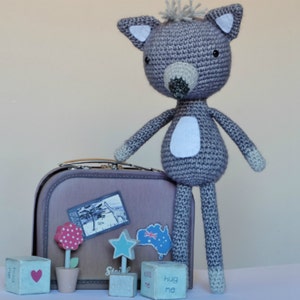 May include: A gray crocheted stuffed animal resembling a wolf or fox, standing next to a brown suitcase with a sticker of Australia on it. The suitcase is surrounded by small wooden blocks with words like "kiss me" and "hug me".