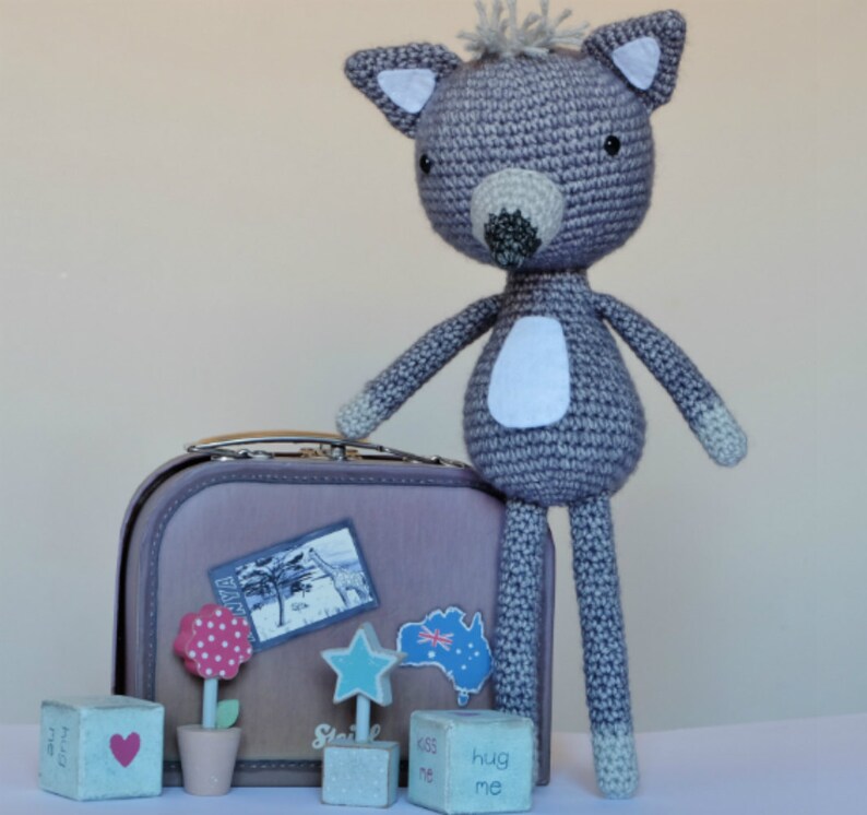 May include: A gray crocheted stuffed animal resembling a wolf or fox, standing next to a brown suitcase with a sticker of Australia on it. The suitcase is surrounded by small wooden blocks with words like "kiss me" and "hug me".