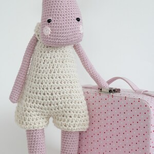 May include: A pink crocheted moose toy wearing a white crocheted outfit, standing next to a pink polka dot suitcase.