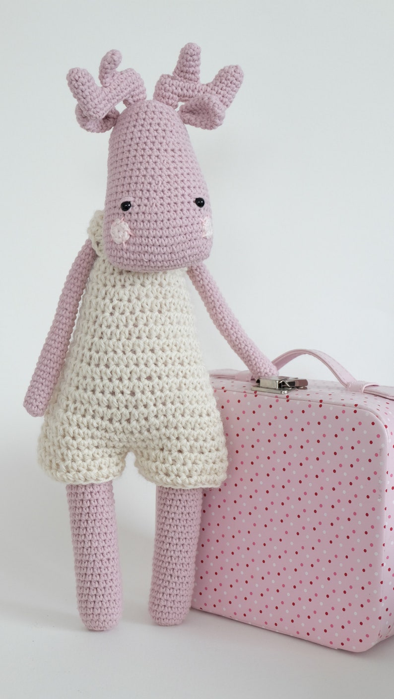 May include: A pink crocheted moose toy wearing a white crocheted outfit, standing next to a pink polka dot suitcase.