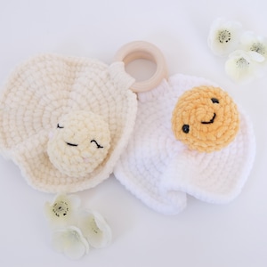 Puede incluir: Un juguete de crochet con forma de huevo frito con una cara sonriente. El huevo está unido a un anillo de madera. El juguete está hecho con hilo blanco y amarillo.