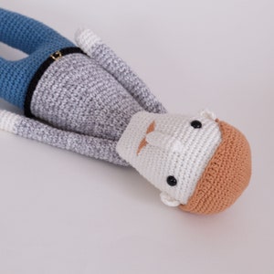 May include: A crocheted stuffed toy person with a gray and white shirt, blue pants, and a brown hat. The toy has black eyes and a white mustache.
