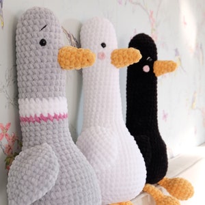 May include: Three crocheted stuffed animal geese. The geese are gray, white, and black. The geese have orange beaks and are sitting on a white surface.