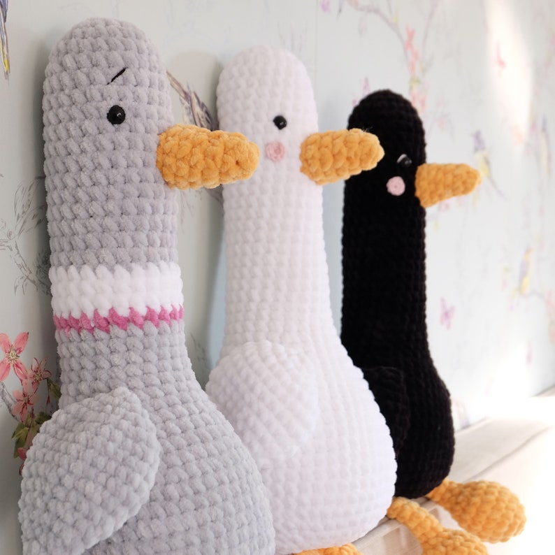 May include: Three crocheted stuffed animal geese. The geese are gray, white, and black. The geese have orange beaks and are sitting on a white surface.