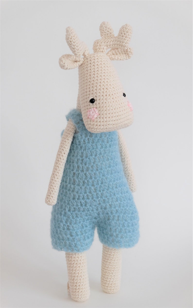 May include: A crocheted stuffed moose toy wearing blue overalls. The moose has a cream-colored body, black eyes, and pink cheeks.