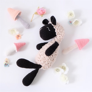 Op de afbeelding: Een gehaakt knuffeldier in de vorm van een schaap met zwarte poten en een zwart gezicht, liggend op een witte achtergrond met roze en witte gehaakte paddenstoelen en bloemen.