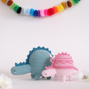 May include: Two crocheted dinosaur toys, one blue and one pink, with a rainbow pom-pom garland in the background.