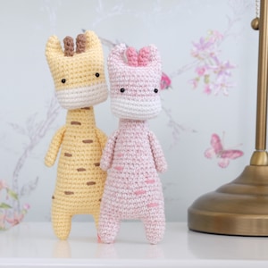 Puede incluir: Dos animales de peluche tejidos a crochet, una jirafa amarilla y un cerdo rosa, de pie uno al lado del otro sobre una superficie blanca.
