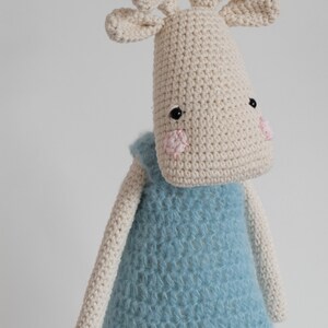 May include: A crocheted stuffed animal in the shape of a moose wearing a blue knitted jumpsuit. The moose has a white body with black eyes and pink cheeks.