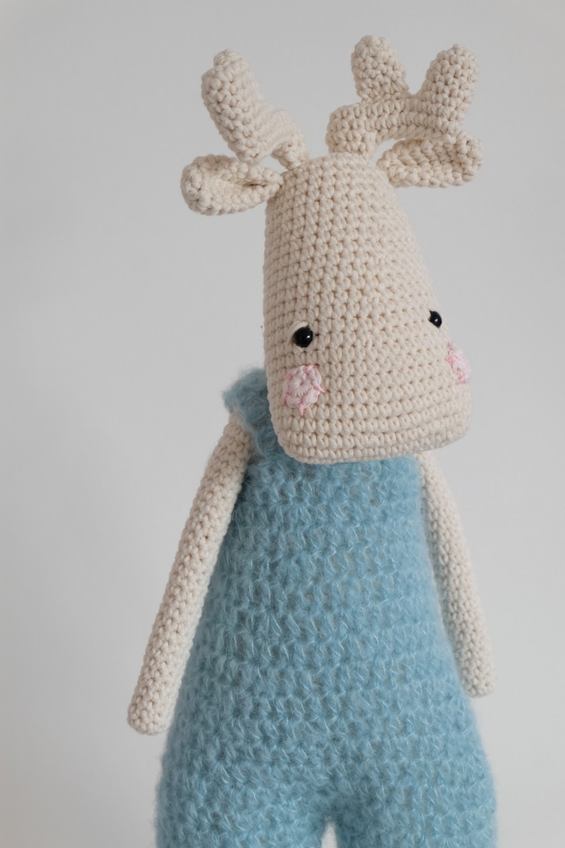 May include: A crocheted stuffed animal in the shape of a moose wearing a blue knitted jumpsuit. The moose has a white body with black eyes and pink cheeks.