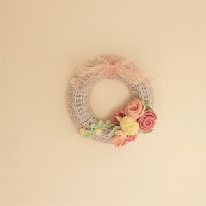 Puede incluir: Una corona de ganchillo hecha a mano en un suave color gris, adornada con flores de ganchillo rosas y amarillas y hojas verdes. Un delicado lazo de cinta rosa se encuentra en la parte superior. Esta corona decorativa es perfecta para a&ntilde;adir un toque artesanal.