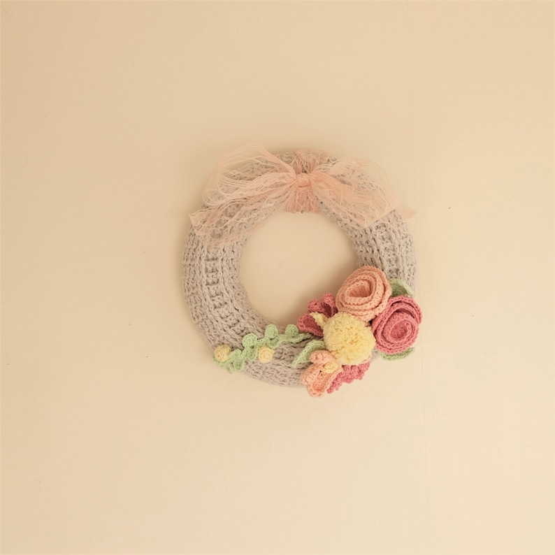 Puede incluir: Una corona de ganchillo hecha a mano en un suave color gris, adornada con flores de ganchillo rosas y amarillas y hojas verdes. Un delicado lazo de cinta rosa se encuentra en la parte superior. Esta corona decorativa es perfecta para a&ntilde;adir un toque artesanal.