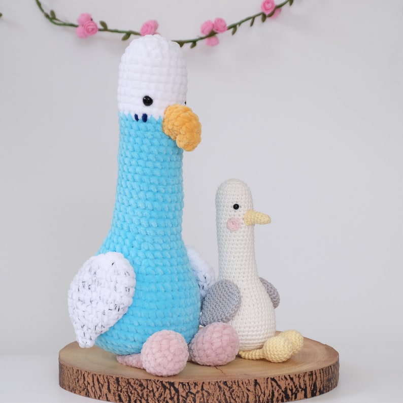 May include: Two crocheted stuffed birds, one blue and one white, sitting on a wooden base. The blue bird has a white beak and yellow accents. The white bird has a pink beak and yellow accents.